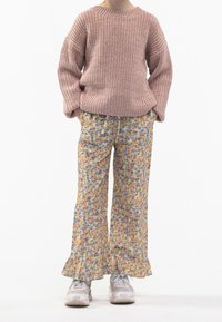 Lapsi, joka on pukeutunut vaaleanpunaiseen paksuun neulepaitaan, keltaisiin kukallisiin leveälahkeisiin housuihin, joissa on röyhelöreunat, ja valkoisiin lenkkitossuihin, seisoo kädet taskuissa.