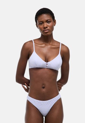Femme aux cheveux courts portant un bikini violet clair texturé avec des bretelles fines et des boutons sur le devant, debout devant un fond clair uni.