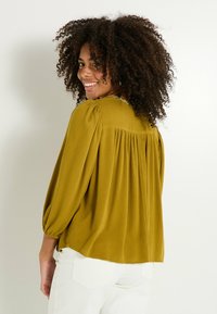 Blouse jaune moutarde avec un dos froncé, des manches longues bouffantes et un décolleté arrondi. Texture lisse avec des détails de couture subtils le long du cou.
