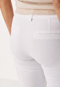 Persoană purtând pantaloni albi mulați cu buzunare aplicate la spate și un top bej din tricot, vizibili de la talie în jos.