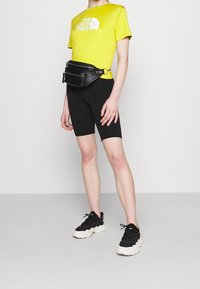 Żółta koszulka z krótkim rękawem z logo "THE NORTH FACE", połączona z czarnymi szortami rowerowymi i czarną saszetką. Czarne sneakersy z białymi podeszwami.