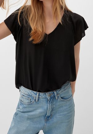 Femme portant un chemisier noir à manches courtes et col en V, rentré dans un jean taille haute bleu clair, sur un fond uni.