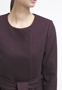 Vínová šatová robota z vlny s rovnou siluetou, kulatým výstřihem a látkovým páskem. Disponuje strukturovanými rameny a minimálními švy.