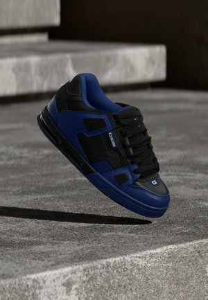 Czarny i niebieski sneaker z sznurowadłami unoszący się nad betonową powierzchnią, w tle kamienne schody.