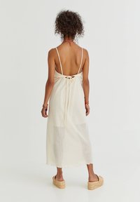 Vestido maxi en crema con un patrón de red texturizado, tirantes ajustables y un detalle de lazo en la parte posterior, combinado con sandalias de esparto.