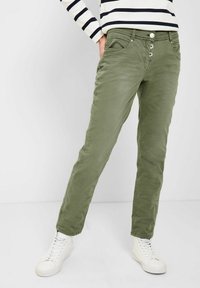 Pantalon vert olive à coupe slim, doté d'une fermeture à trois boutons, de poches avant et d'une texture lisse. Associé à des baskets blanches.