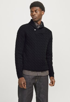 Schwarzer Strickpullover mit Schalkragen, ausgestattet mit einem einzigen Knopfverschluss. Über einem gestreiften Hemd getragen und kombiniert mit dunklen Jeans.