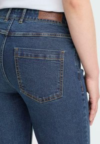 Tumši zilie džinsi ar klasisku dizainu, kas aprīkoti ar ādas etiķeti, divām aizmugurējām kabatām un oranžām šuvēm.