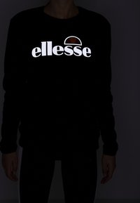 Sudadera negra de manga larga con un destacado logo reflectante en blanco de "ellesse" y un diseño marrón semicircular encima del texto.