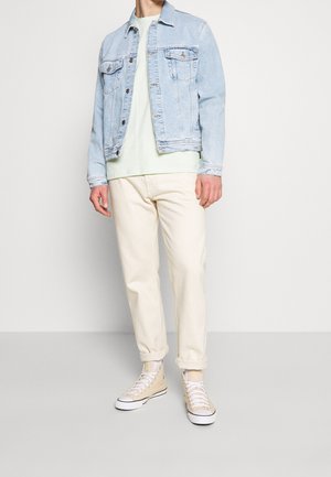 Mann trägt eine hellblaue Jeansjacke, ein blassgrünes Hemd, elfenbeinfarbene Hose und beigen High-Top-Sneaker, steht vor weißem Hintergrund.