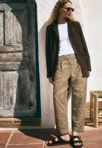 Brauner Wildlederblazer über einem weißen Oberteil, kombiniert mit lockeren, gemusterten khakihosen und schwarzen Sandalen. Minimalistisches Design und natürliche Texturen.