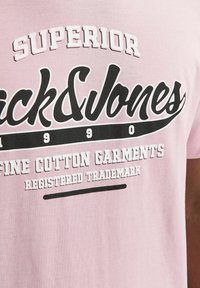 Rosa Baumwoll-T-Shirt mit auffälligem schwarzen "Jack&Jones"-Logo und weißem Text, der "SUPERIOR" und "FEINE BAUMWOLLBEKLEIDUNG" umfasst.