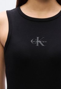 Μαύρο ριπ τοπ χωρίς μανίκια με στρογγυλή λαιμόκοψη, που φέρει διακριτικό ασημένιο ανάγλυφο λογότυπο "Calvin Klein" στο κέντρο της περιοχής του στήθους.
