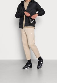 Zwarte zip-up jas, beige sweatshirt, beige tapered broek met Nike-logo, zwarte Nike sneakers en een kleine zwarte tas met witte accenten.