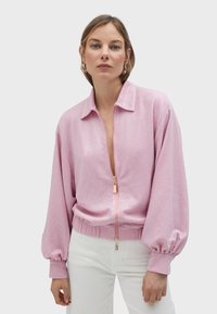 Ružičasta jakna s patentnim zatvaračem i teksturiranim uzorkom, s ovratnikom i balonskim rukavima. Ima zlatni patentni zatvarač i rebrasti rub. Nosi se preko bijelih hlača.