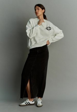 Felpa bianca con logo grafico nero, abbinata a una gonna lunga in denim nera con spacco frontale e sneakers bianche con strisce nere.