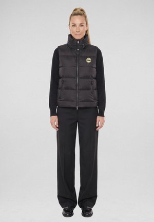 Vrouw met blond haar in een paardenstaart draagt een zwarte gewatteerde vest met rits en zwarte wijde broek, staand voor een effen grijze achtergrond.
