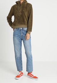 Olivgrön fleecepullover med halv dragkedja, framfickor och huva, tillsammans med ljusblå raka jeans och orange låga sneakers.