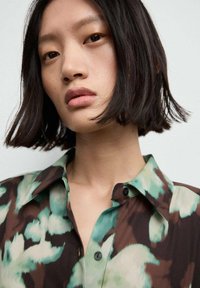 Camicia con un motivo floreale in tonalità di verde e marrone, con un design a colletto, bottoni e un tessuto liscio e leggero.