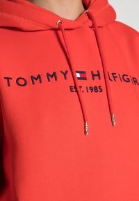 Röd bomullshuvtröja med dragsko, prydd med broderad "TOMMY HILFIGER"-text och logotyp i marinblått och vitt. Guldiga detaljer.