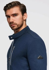 Marinblå strukturerad jacka med ståkrage, utrustad med en silverfärgad dragkedja, bröstficka och en svart triangulär logotyp patch på ärmen.