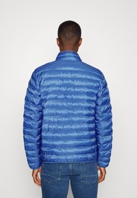 Blaue Steppjacke mit einer gesteppten Optik, ausgestattet mit einem Stehkragen und einer glatten Textur. Die Jacke wird über dunkelblauen Jeans getragen.