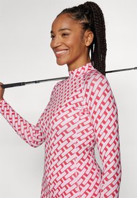 Pikkade varrukatega, kehaümbermoodustav golfipluus valges, millel on punane geomeetriline muster. Kõrge krae ja sile, veniv kangas. Golfikepp resting õlal.