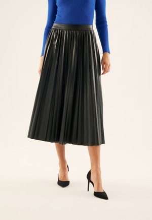 Vrouw die een zwarte geplooide midirok, een blauw shirt met lange mouwen en zwarte hoge hakken met puntige neus draagt, staand tegen een effen achtergrond.
