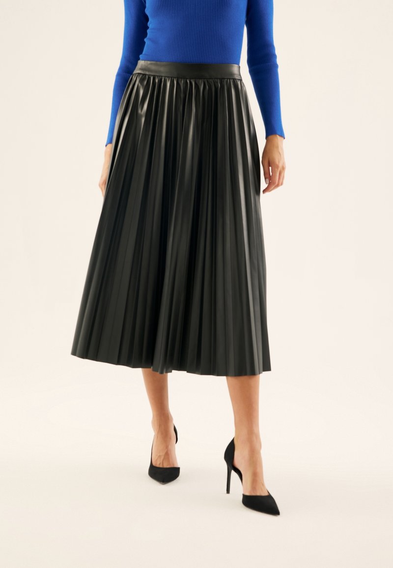 Femme portant une jupe midi plissée noire, un haut bleu à manches longues et des escarpins noirs à bouts pointus, debout devant un fond uni.