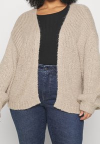 Beige gebreid vest met verlaatte schouders, open voorkant, geribbelde textuur en lange, brede mouwen, gecombineerd met een zwart topje en blauwe spijkerbroek.