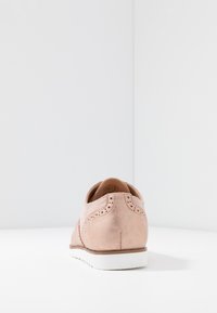 Vaaleanpunainen nahkakenkä, jossa on brogue-yksityiskohtia, sileä pinta, valkoinen kumipohja ja pyöreä kärki. Näkymä takaa.