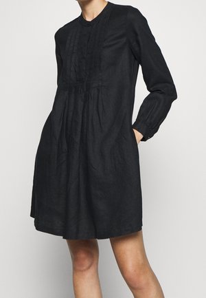 Woman wearing a knee-length black long-sleeve dress with buttoned cuffs and vertical pleats on the chest, posing with hands in pockets.