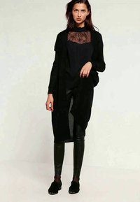 Frau in schwarzem Spitzentop, langem schwarzen Cardigan, schwarzen Lederhosen und schwarzen Schuhen, die vor einem schlichten hellen Hintergrund steht.