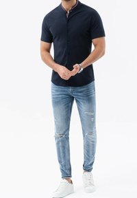 Camisa de manga corta azul marino con cuello recto y cierre de botones, combinada con jeans claros rasgados y zapatillas blancas.