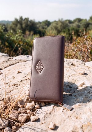 Una cartera Replay de cuero marrón está de pie sobre una superficie rocosa iluminada por el sol, con hierbas secas y árboles verdes al fondo.