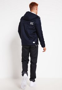 Giacca blu navy con cappuccio e chiusura con zip, logo bianco sulla schiena e jeans neri. Il modello indossa sneakers bianche ed è rivolto di spalle.