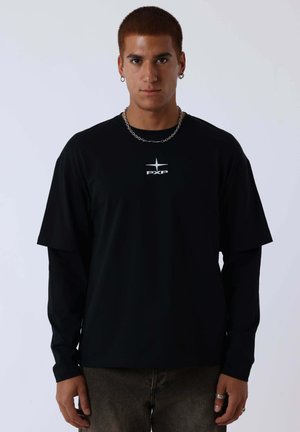 Giovane uomo che indossa magliette nere a maniche lunghe e corte sovrapposte con una collana a catena argentata e piccoli orecchini a cerchio su uno sfondo semplice.