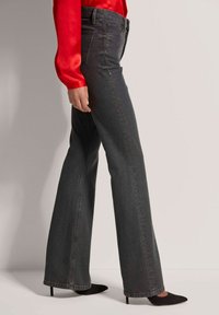 Schwarze Denim-Jeans mit Schlag und hoher Taille, featuring dezente Steppnähte, getragen mit einer roten Satinbluse und schwarzen spitzen Absätzen.
