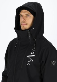 Schwarze Kapuzenjacke mit hohem Kragen, frontalem Reißverschluss und Kordelzügen. Besitzt ein weißes Logo auf der Brust und einen Aufnäher am Ärmel.