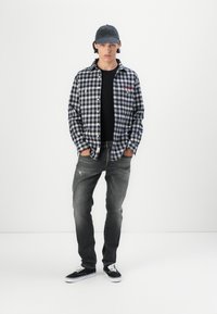 Zwart-wit geruite shirt, zwart T-shirt, grijze spijkerbroek en zwarte sneakers. Pet met logo. Staand tegen een effen witte achtergrond.