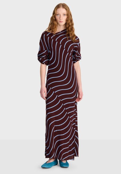 Femme aux longs cheveux ondulés portant une longue robe sombre avec des rayures ondulées claires et des chaussons bleus, debout devant un fond uni.