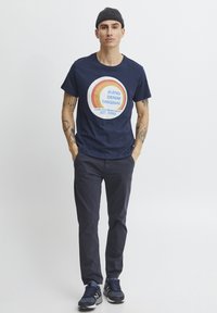 Jūrų mėlynos medvilnės marškinėliai su apvaliu grafiniu dizainu, kuriame yra tekstas mėlyna ir oranžine spalvomis, derinami su tamsiomis kelnėmis ir jūrų mėlynais sportiniais batais.