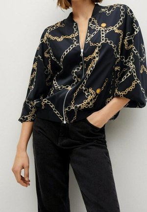 Vrouw draagt een zwarte blouse met ritssluiting en print van gouden en zilveren kettingen, gecombineerd met hoogtalige zwarte broek, hand in de zak.