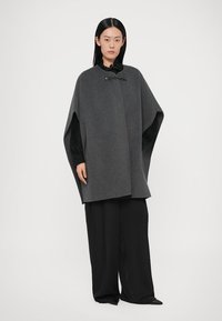 Siva vunena kapa opuštenog kroja s prednjim zatvaračem na kopču i širokim rukavima, uparena s crnim hlačama širokih nogavica.