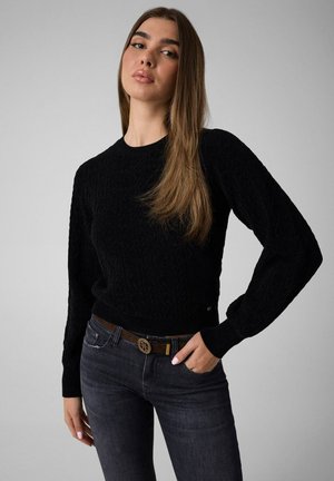 Jeune femme aux longs cheveux raides portant un pull texturé noir, un jean foncé et une ceinture marron avec une boucle décorative.