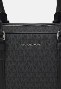 Sac à main noir en tissu texturé, arborant le motif "MK", avec des détails en métal argenté et une fermeture éclair. Logo affiché de manière proéminente à l'avant.