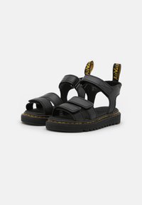 Black sandals with a chunky sole, featuring wide adjustable straps, open toe design, and yellow stitching along the edge, reinforced with tags.