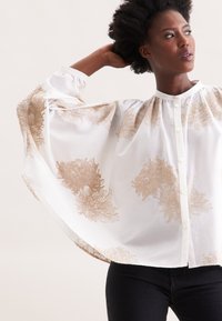Blusa bianca con ricami floreali dorati, vestibilità ampia, colletto alto, chiusura con bottoni e maniche morbide, abbinata a pantaloni neri.