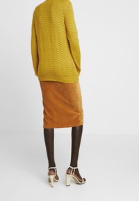 Musztardowy, fakturowany sweter z zygzakowatym wzorem, połączony z brązową spódnicą ołówkową z kordurowego materiału i beżowymi sandałami na obcasie.