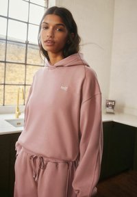 Hele roosa kapuutsiga särk valge "self." logoga, millel on mugav lõige, nööridega vöökoht ja pehme kangas. Stylenud koos sobivate jogger-pükstega.